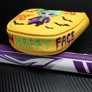 May include: Yellow golf club head cover with an embroidered Joker design and the words "ON HAPPY FACE". The cover also features embroidered bats. The golf club handle is purple and white.