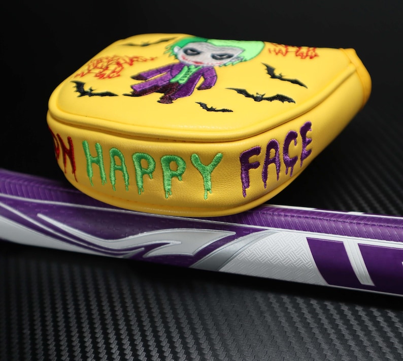 May include: Yellow golf club head cover with an embroidered Joker design and the words "ON HAPPY FACE". The cover also features embroidered bats. The golf club handle is purple and white.
