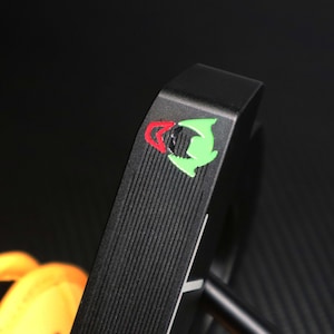 May include: A black golf putter with a textured face and a colorful logo featuring red and green accents. The putter's design includes a straight shaft and a rounded head, suitable for golf enthusiasts.