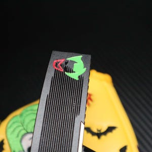 May include: Black and green Joker-themed accessory with a ridged surface. The accessory features a red and green graphic of the Joker's face. A yellow object with a Joker graphic and bat silhouettes is visible in the background.