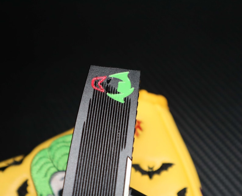 May include: Black and green Joker-themed accessory with a ridged surface. The accessory features a red and green graphic of the Joker's face. A yellow object with a Joker graphic and bat silhouettes is visible in the background.