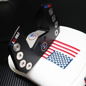 May include: Black metal golf club head cover with the text "MEZZ.MAX" and "L.A. GOLF". The cover is white with an embroidered American flag and the word "UNITY".