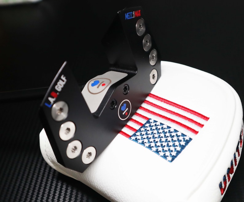 May include: Black metal golf club head cover with the text "MEZZ.MAX" and "L.A. GOLF". The cover is white with an embroidered American flag and the word "UNITY".