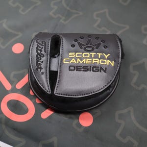 May include: A black golf putter cover with the Titleist logo on the side. The cover features the words "SCOTTY CAMERON DESIGN" in gold lettering, along with a crown design. The cover is made of a textured material.