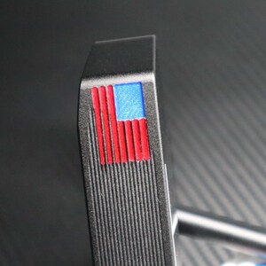 May include: A black golf putter head with a textured surface. The putter features a small American flag design in red, white, and blue on the top. The putter's shaft is visible in the background.