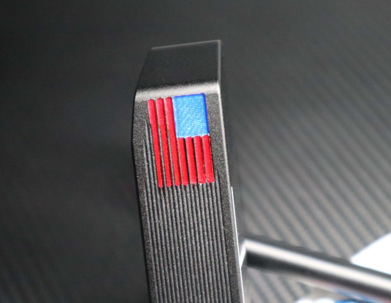 May include: A black golf putter head with a textured surface. The putter features a small American flag design in red, white, and blue on the top. The putter's shaft is visible in the background.