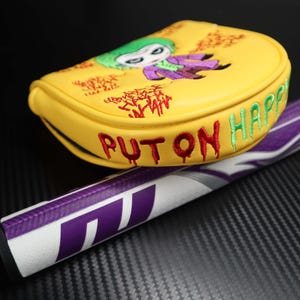 May include: Yellow golf club head cover with an embroidered Joker design, green hair and purple suit. The cover has the words "PUT ON HAPPY" in red and green. A purple and white golf club handle is visible.