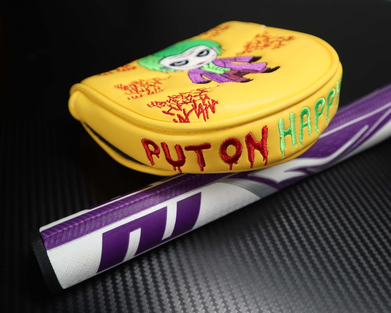 May include: Yellow golf club head cover with an embroidered Joker design, green hair and purple suit. The cover has the words "PUT ON HAPPY" in red and green. A purple and white golf club handle is visible.
