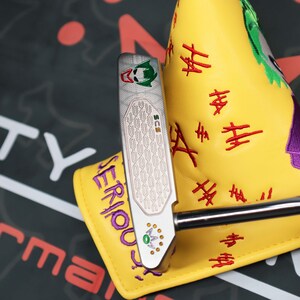 May include: A silver golf putter with a Joker-themed design, featuring a green and red logo and a chain pattern on the face. The putter is paired with a yellow headcover embroidered with "SERIOUS!" and "HA HA" in red.