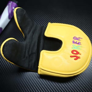 May include: Yellow and black golf putter cover with the word "JOKER" embroidered in red, green, and purple. The cover has a U-shaped design and a black interior. A purple and white golf club grip is visible in the background.