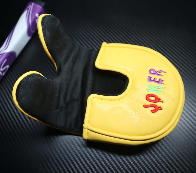 May include: Yellow and black golf putter cover with the word "JOKER" embroidered in red, green, and purple. The cover has a U-shaped design and a black interior. A purple and white golf club grip is visible in the background.