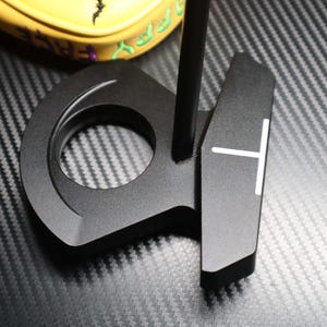 May include: A black golf putter with a circular design and a white T-shaped alignment aid. The putter is made of metal and has a matte finish. A yellow golf club cover with green text is visible in the background.