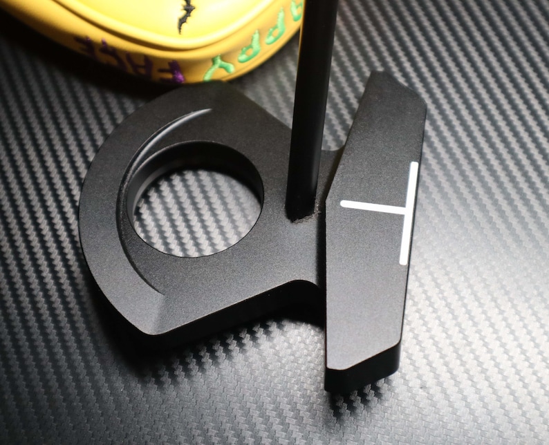 May include: A black golf putter with a circular design and a white T-shaped alignment aid. The putter is made of metal and has a matte finish. A yellow golf club cover with green text is visible in the background.