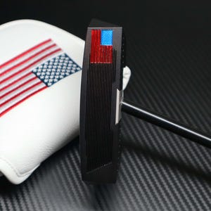 May include: A black golf putter with a red, white, and blue American flag design on the top. The putter has a black shaft and a white head cover with an embroidered American flag.