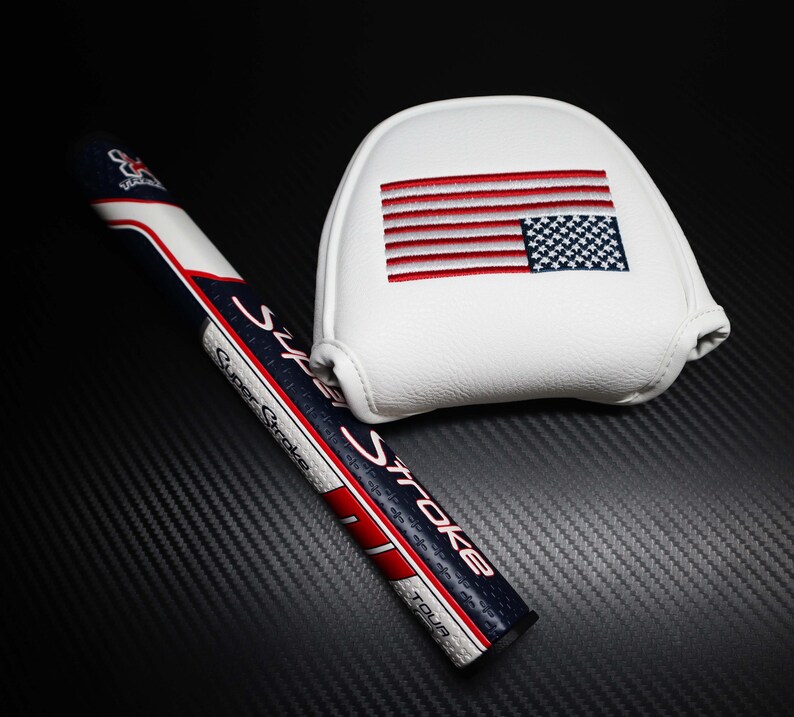 May include: A golf putter grip and head cover set. The grip is navy blue, white, and red with the text "SuperStroke". The white head cover features an embroidered American flag.
