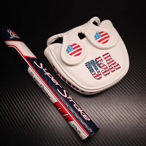 May include: A white golf putter grip and head cover set. The grip is navy blue, white, and red with the text "Super Stroke". The head cover is white with the text "USA" and American flag designs.