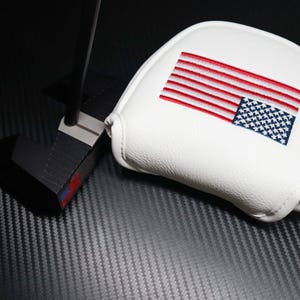 May include: A black golf putter with a white head cover. The head cover features an embroidered American flag in red, white, and blue. The putter is set against a dark, textured background.