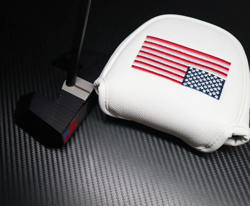 May include: A black golf putter with a white head cover. The head cover features an embroidered American flag in red, white, and blue. The putter is set against a dark, textured background.
