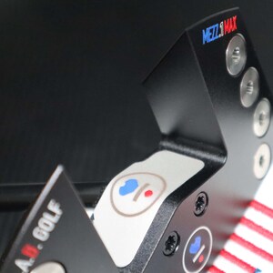May include: Black golf putter with the text "MEZZ.1 MAX" in blue and red. The putter has a white section with blue and red circular graphics. The putter is made of metal and has silver screws.
