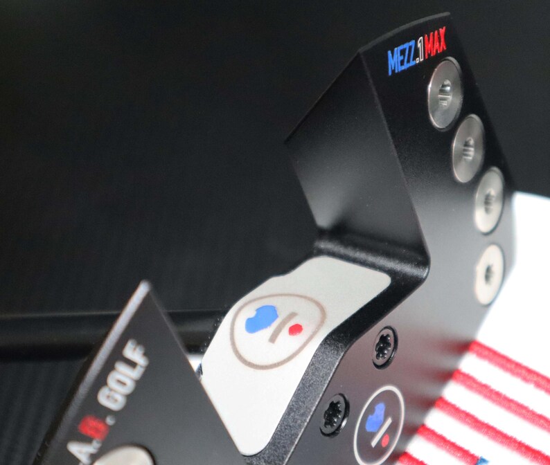 May include: Black golf putter with the text "MEZZ.1 MAX" in blue and red. The putter has a white section with blue and red circular graphics. The putter is made of metal and has silver screws.