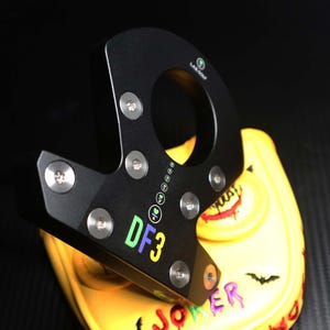 May include: Black golf putter head with the text "DF3" in green, yellow, and purple. The putter is resting on a yellow golf club head cover with a Joker-themed design. The cover features a smiling face, a bat, and the word "JOKER".