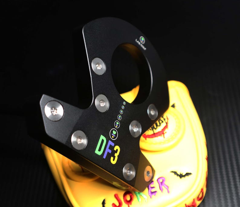 May include: Black golf putter head with the text "DF3" in green, yellow, and purple. The putter is resting on a yellow golf club head cover with a Joker-themed design. The cover features a smiling face, a bat, and the word "JOKER".