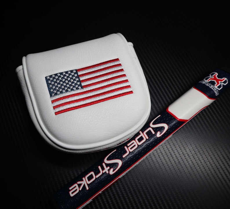 May include: White golf putter cover with an embroidered American flag in red, white, and blue. A golf club grip with the text "Super Stroke" in white and red is also visible.