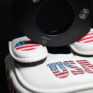May include: White golf club head covers with embroidered American flag designs and the word "USA". A black golf club is positioned above the covers. The covers are made of a soft, textured material.