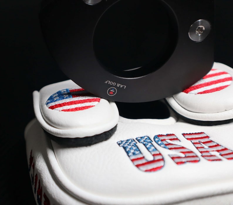 May include: White golf club head covers with embroidered American flag designs and the word "USA". A black golf club is positioned above the covers. The covers are made of a soft, textured material.