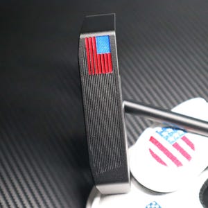 May include: A black golf putter with a red, white, and blue American flag design on the top. The putter has a grooved face and a black shaft. The putter is resting on a white surface with a matching flag design.