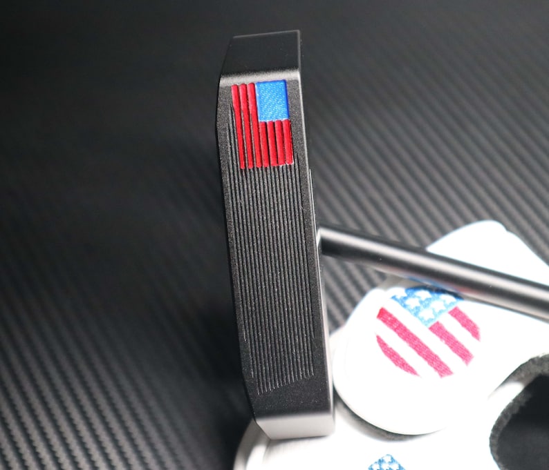 May include: A black golf putter with a red, white, and blue American flag design on the top. The putter has a grooved face and a black shaft. The putter is resting on a white surface with a matching flag design.