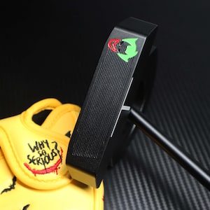 May include: Black golf putter with a green and red logo. The putter is positioned next to a yellow golf glove with the words "WHY SO SERIOUS?" embroidered in red and white.