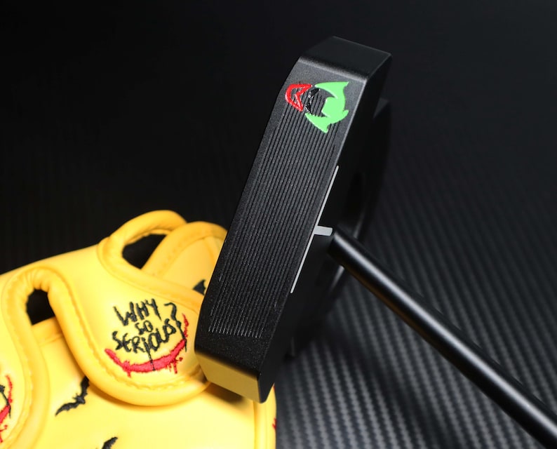 May include: Black golf putter with a green and red logo. The putter is positioned next to a yellow golf glove with the words "WHY SO SERIOUS?" embroidered in red and white.
