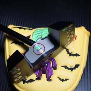 May include: A black golf putter with a yellow head cover featuring a cartoon Joker, green leaves, and black bats. The putter has a gray top with a green and yellow symbol. The putter is designed for the golf course.