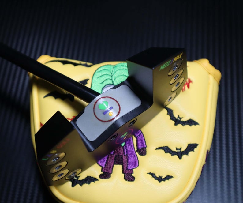 May include: A black golf putter with a yellow head cover featuring a cartoon Joker, green leaves, and black bats. The putter has a gray top with a green and yellow symbol. The putter is designed for the golf course.