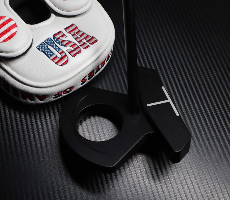 May include: A black golf putter with a white line design and a matching head cover. The head cover is white with the word "USA" in red, white, and blue, and the words "STATES OF AMERICA" in red.