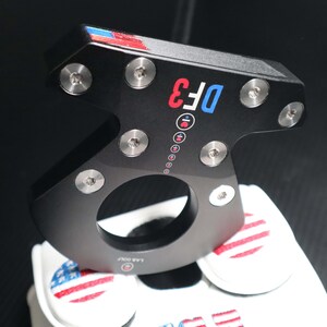 May include: Black golf putter with the text "DF3" in red and blue. The putter has silver screws and a circular opening. The top has red, white, and blue accents. A white golf glove with an American flag design is visible.