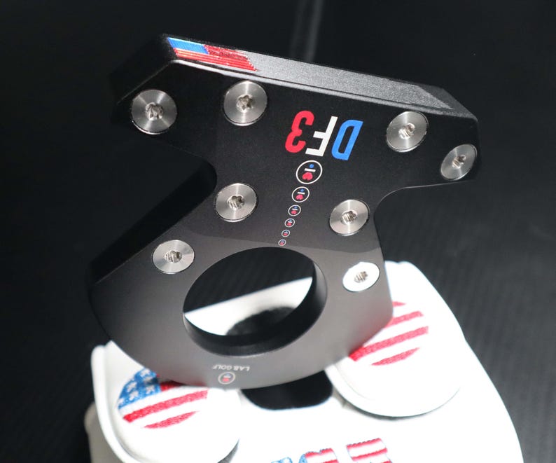 May include: Black golf putter with the text "DF3" in red and blue. The putter has silver screws and a circular opening. The top has red, white, and blue accents. A white golf glove with an American flag design is visible.