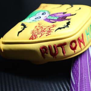 May include: Yellow golf club head cover with an embroidered image of the Joker character. The cover has the words "PUT ON HAHA" and "HAPPY" embroidered in red and green. Black bats and other graphics are also embroidered on the cover.