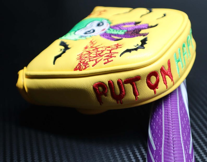 May include: Yellow golf club head cover with an embroidered image of the Joker character. The cover has the words "PUT ON HAHA" and "HAPPY" embroidered in red and green. Black bats and other graphics are also embroidered on the cover.
