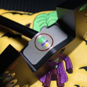 May include: A black golf putter with the text "MEZZ MAX" on the side. The putter has a grey top with a circular design. The putter is resting on a yellow golf club cover with a Joker design and black bat silhouettes.