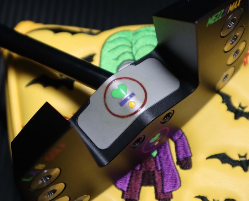 May include: A black golf putter with the text "MEZZ MAX" on the side. The putter has a grey top with a circular design. The putter is resting on a yellow golf club cover with a Joker design and black bat silhouettes.