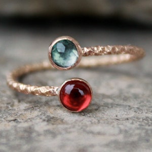 以下が含まれることがあります： テクスチャ加工されたバンドと2つの宝石が付いた金のリング。大きな宝石は濃い赤色で、小さな宝石は明るい緑色です。