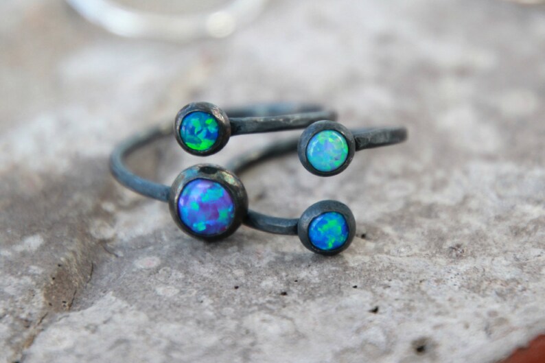 Puede incluir: Primer plano de un anillo de plata con cuatro piedras de &oacute;palo iridiscentes. El anillo tiene un dise&ntilde;o &uacute;nico con las piedras engastadas en biseles. Los &oacute;palos muestran tonos de azul, verde y morado.