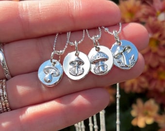 May include: Four silver mushroom pendant necklaces. Each pendant features a different mushroom design on a round disc. The necklaces are displayed against a blurred background of orange and brown flowers.
