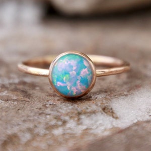 May include: A delicate gold ring with a round, light blue opal gemstone. The ring is set on a textured, brown surface.