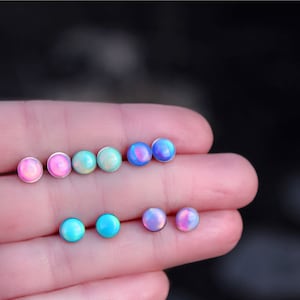 May include: A set of eight iridescent opal stud earrings in various shades of pink, blue, and green. The earrings are displayed on a hand.