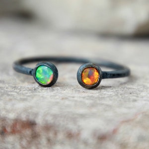 Puede incluir: Anillo abierto con una banda de metal oscuro. El anillo presenta dos piedras de &oacute;palo engastadas, una con tonos verdes y azules, la otra con tonos naranjas y amarillos. El anillo est&aacute; sobre un fondo neutro y texturizado.
