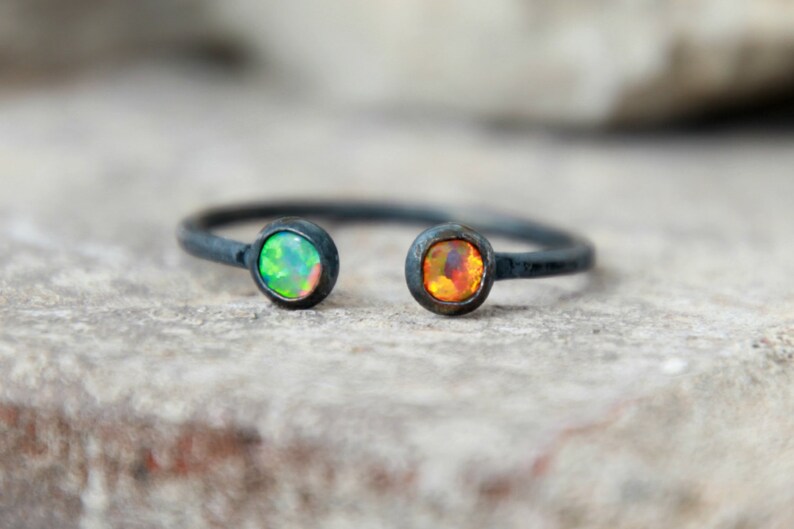 Puede incluir: Anillo abierto con una banda de metal oscuro. El anillo presenta dos piedras de &oacute;palo engastadas, una con tonos verdes y azules, la otra con tonos naranjas y amarillos. El anillo est&aacute; sobre un fondo neutro y texturizado.