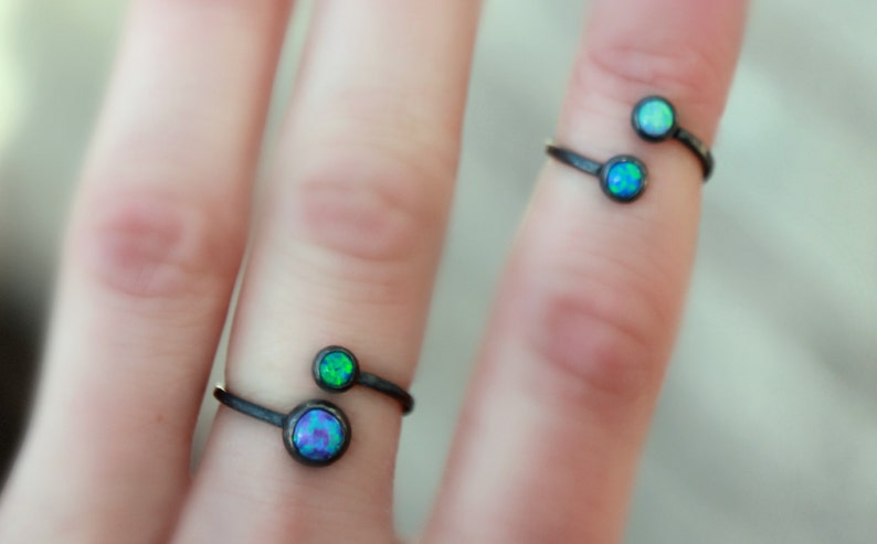 Puede incluir: Dos anillos abiertos con un acabado de metal oscuro, cada uno con dos piedras iridiscentes tipo &oacute;palo. Las piedras muestran tonos de azul y verde. Los anillos se muestran en un dedo.
